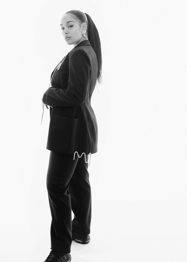 Monochrome portrait of a stylish adult woman in a black blazer and trousers, with her hair pulled back in a high ponytail, against a white backdrop.