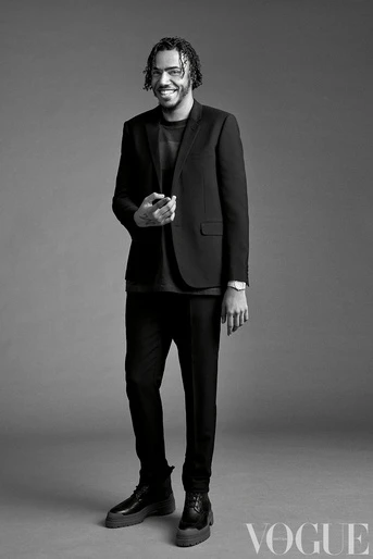 Black and white portrait of a smiling adult male in a blazer, t-shirt, pants and lug-sole boots, standing in a studio against a solid grey background.