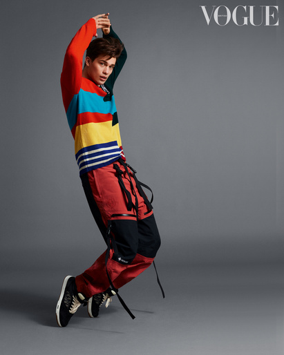Young man strikes fashion pose indoors in colorful striped sweater, red and black pants, and black shoes against a gray backdrop.
