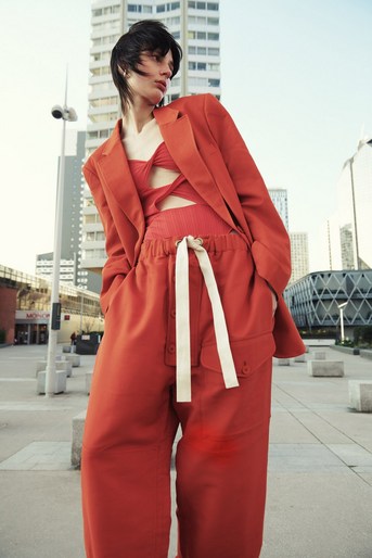 Fashionable woman posing outdoors wearing an orange suit jacket and pants with a matching strappy top, against a modern city backdrop.