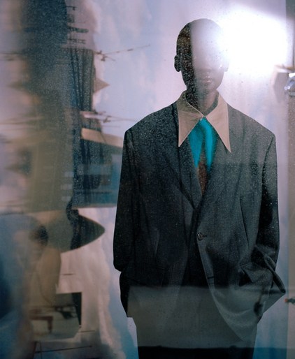 An African American man stands wearing a suit with a teal shirt and tie, reflected in a window with blurred urban background, and sunlight.