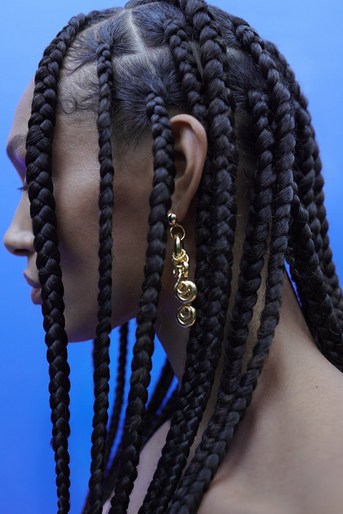 Close-up profile of a woman with black braided hair and dangling gold earrings against a bright blue background, showcasing beauty and style.