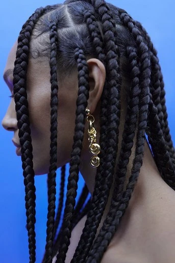 Close-up profile of a woman with black braided hair and dangling gold earrings against a bright blue background, showcasing beauty and style.