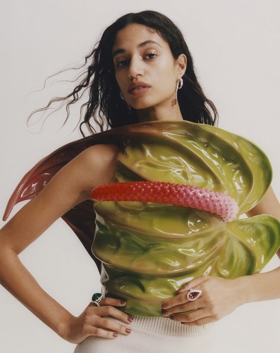 Elegant woman with long dark hair poses in front of a white backdrop wearing a green floral top and jewelry with rings.
