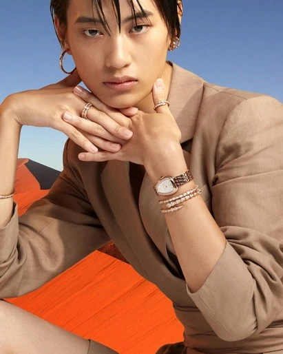 Person wearing jewelry and tan blazer jacket in a studio shot with a blue sky and tan color gradient in the background.