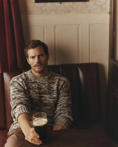 A man with fair skin and brown hair sits inside a bar booth holding a glass of dark beer with a white foam head. He wears a knit sweater.