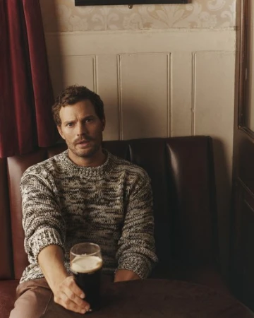 A man with fair skin and brown hair sits inside a bar booth holding a glass of dark beer with a white foam head. He wears a knit sweater.