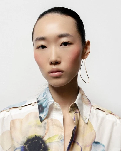 Close-up portrait of an Asian woman with slicked-back hair wearing a flower-print shirt and a delicate, curved earring, set against a white background.