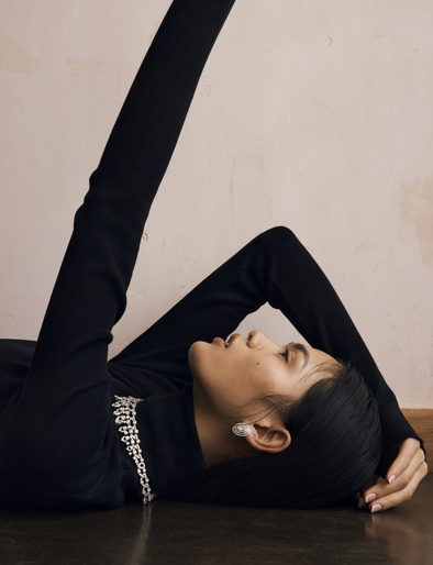 A woman in a black turtleneck with diamond jewelry poses while lying on the floor in front of a pinkish-beige wall.