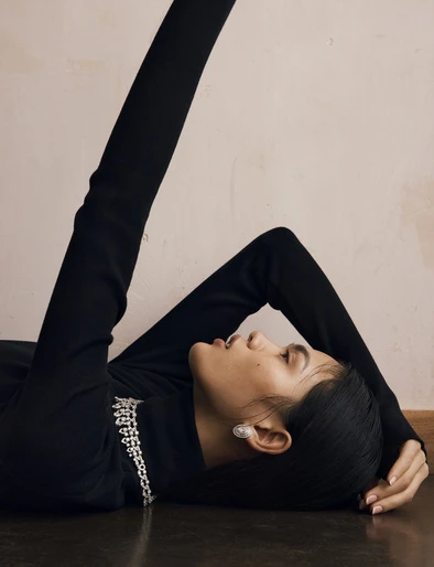 A woman in a black turtleneck with diamond jewelry poses while lying on the floor in front of a pinkish-beige wall.