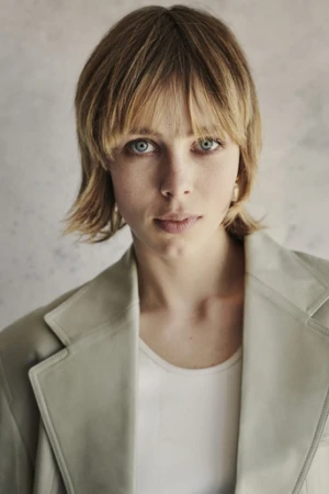 Portrait of a blonde woman with bangs wearing a sage green blazer over a white top against a mottled background.