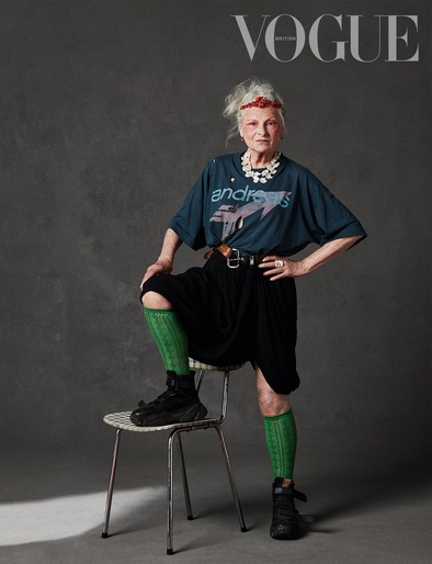 Fashion editorial shot of older woman, Vivienne Westwood, wearing a graphic t-shirt, black skirt, knee socks, and sneakers, sitting on a chair.