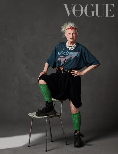 Fashion editorial shot of older woman, Vivienne Westwood, wearing a graphic t-shirt, black skirt, knee socks, and sneakers, sitting on a chair.