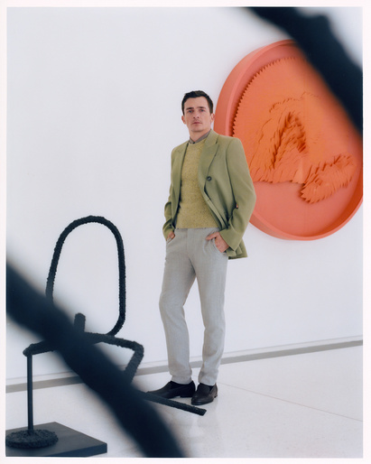 Adult man in a suit jacket and sweater poses in front of modern art pieces in a gallery, hands in his pockets with a neutral expression.