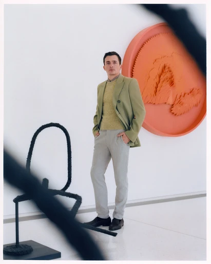 Adult man in a suit jacket and sweater poses in front of modern art pieces in a gallery, hands in his pockets with a neutral expression.