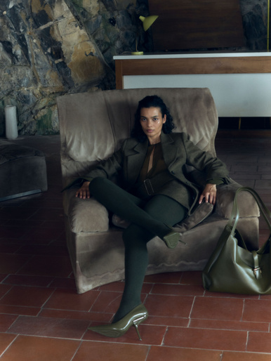 Brunette woman in green business suit seated in armchair with handbag, green heels on red terracotta tile floor, stone wall backdrop.