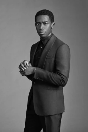 Black man wearing a dark tailored suit and black shirt standing against a seamless grey studio backdrop