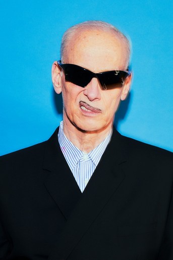 Close-up portrait of an older man with slicked-back hair, wearing sunglasses and a suit, set against a solid blue background.