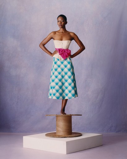 A striking fashion shot of a Black female model in a corset and patterned skirt, balancing on a spool, against a lilac backdrop.