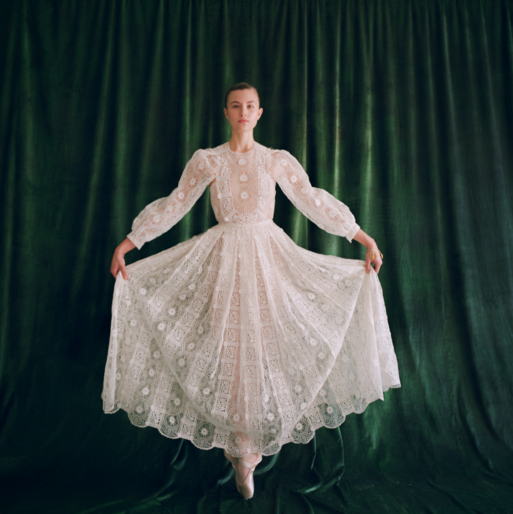 Elegant woman in white lace dress strikes ballet pose in front of dark green velvet backdrop, illuminated by soft studio lighting.
