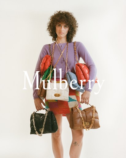 Woman in lilac cardigan poses with multiple quilted leather handbags in various colors against a neutral backdrop in a fashion shot.
