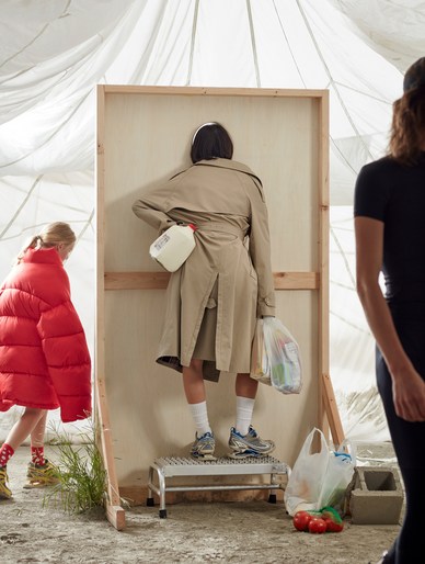 Back view of an adult stands on a step stool with a milk jug in their coat, a child and adult standing nearby, set on a tarp covered ground.