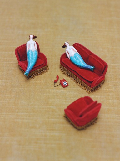 Two mermaid figurines rest on miniature red velvet couches, accompanied by a tiny red rotary telephone, set against a warm beige backdrop.