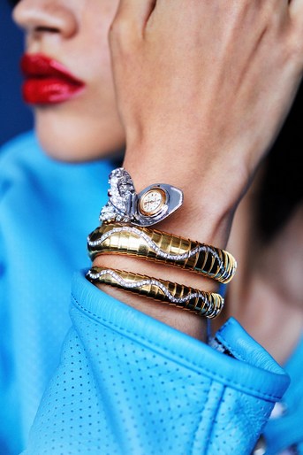 A woman showcases a gold, snake-shaped Bulgari watch with diamond accents against a blue backdrop, emphasizing luxury jewelry details.