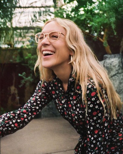 Smiling woman with blonde hair, wearing glasses and a floral dress, stands indoors with lush greenery and a rock wall in the background.