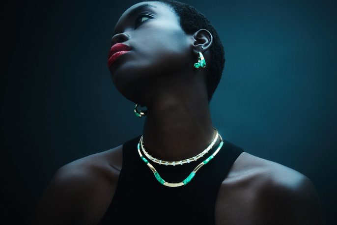 Portrait of a Black woman with red lips wearing jewelry, including a necklace and earrings, looking up against a dark teal background.