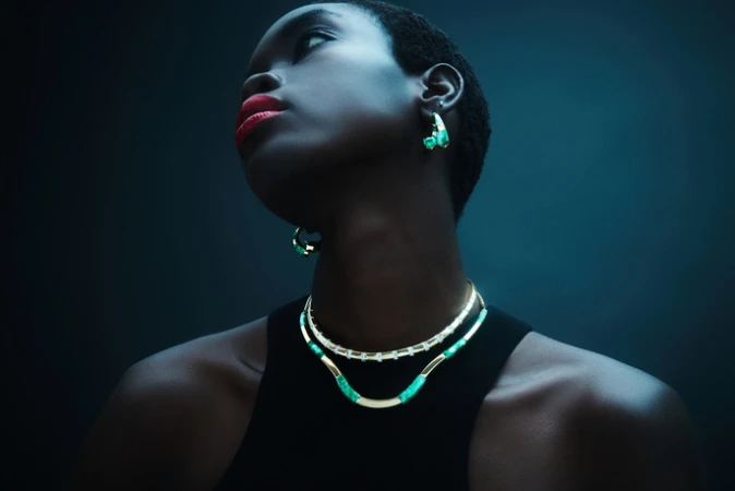 Portrait of a Black woman with red lips wearing jewelry, including a necklace and earrings, looking up against a dark teal background.