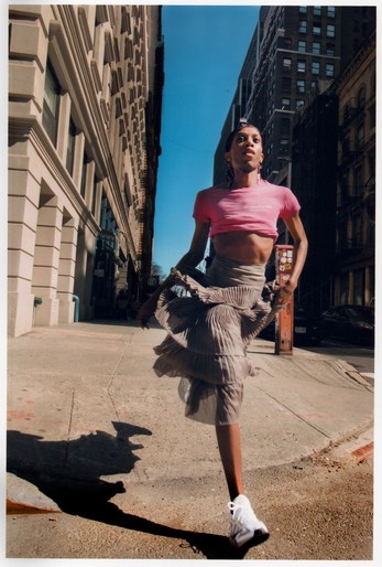 Woman in pink top and flowing skirt runs on city street with buildings and blue sky in background, captured in full shot.