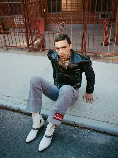 Man sitting on a curb wearing a black leather jacket and boots, with a red metal fence and a brick building in the background.