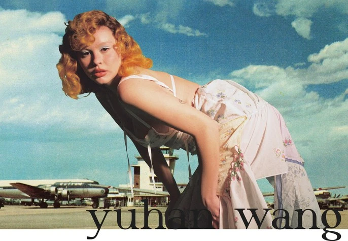 A fashion portrait of an adult female with orange hair in a patterned dress against a backdrop of airplanes and a blue cloudy sky.