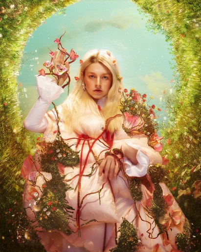 Woman with blonde hair poses in a garden setting, wearing a decorative dress adorned with flowers, standing under a floral archway.