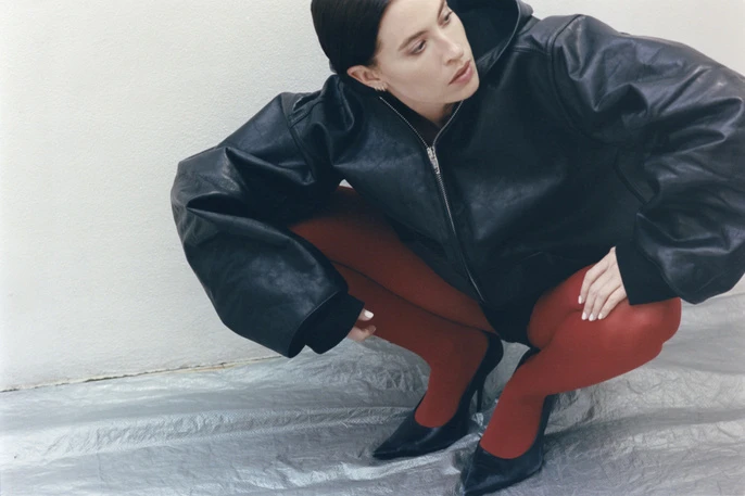 Woman in black leather jacket and red tights squatting on silver surface in studio, looking off to the side in a fashion pose.
