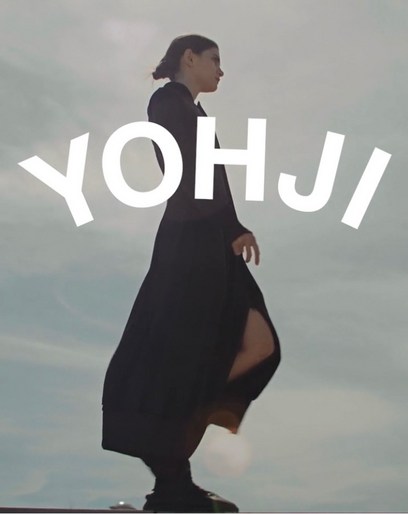 A woman in a dark dress and shoes walks outdoors in front of the white text “YOHJI,” with a cloudy sky and tan ground in the background.