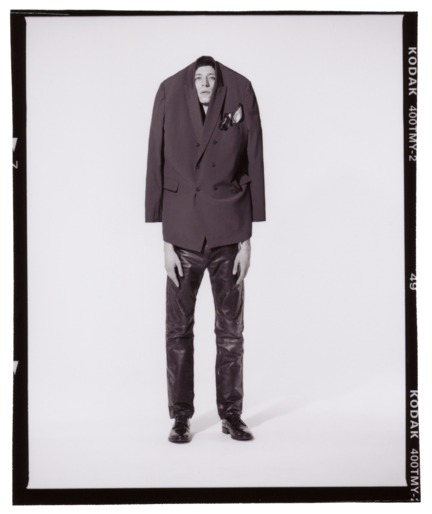 A man in an oversized blazer and leather pants poses in a photo studio, showcasing a blend of high fashion and unconventional style.