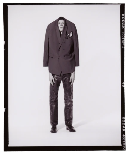 A man in an oversized blazer and leather pants poses in a photo studio, showcasing a blend of high fashion and unconventional style.