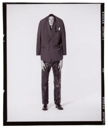 A man in an oversized blazer and leather pants poses in a photo studio, showcasing a blend of high fashion and unconventional style.