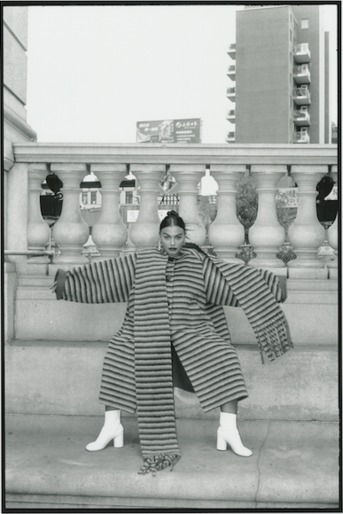 Monochrome fashion shot of a female adult in a striped coat and matching pants, striking a pose in white boots against a building backdrop.