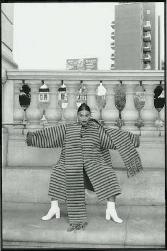 Monochrome fashion shot of a female adult in a striped coat and matching pants, striking a pose in white boots against a building backdrop.