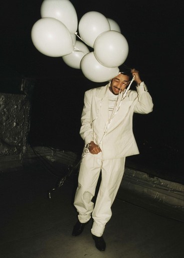 Stylish adult male in a white suit poses indoors while holding a bunch of white balloons, illuminated by a directed light source. 