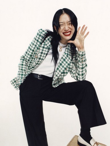 Cheerful Asian model strikes a pose in the studio wearing a patterned blazer, top, and black slacks while standing on a wooden box.