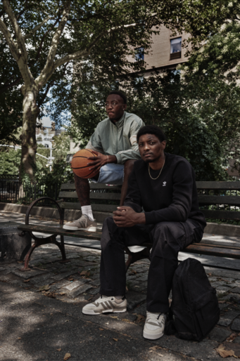 Two adult men sit on a park bench beneath leafy trees. One man holds a basketball. A backpack rests on the ground nearby.