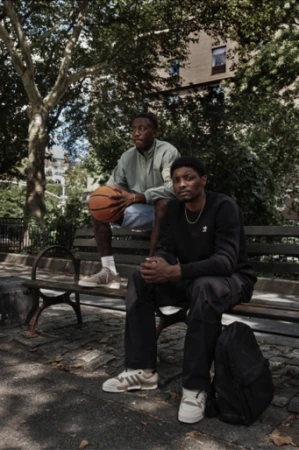 Two adult men sit on a park bench beneath leafy trees. One man holds a basketball. A backpack rests on the ground nearby.