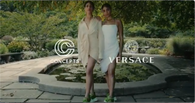Two women pose in designer dresses in a garden setting, with Versace and Concepts logos flanking a pond and greenery, creating a fashion-forward image.