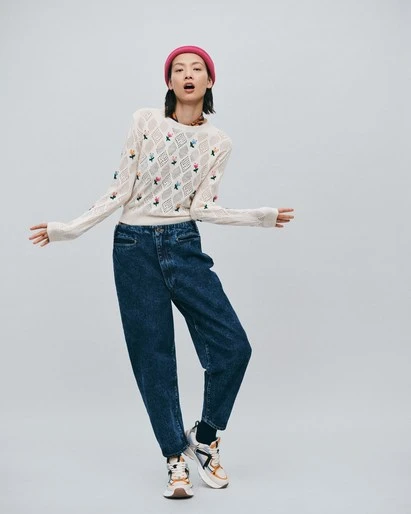 Fashion shot of a young Asian woman wearing a floral knit sweater, dark denim baggy jeans, a pink beanie, and platform sneakers in a studio.