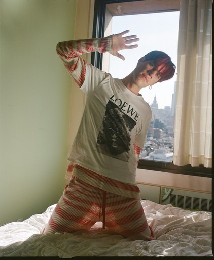 Adult male poses on a bed by a window in an indoor setting, wearing a striped sweater and shorts with a Loewe t-shirt, city in the background.