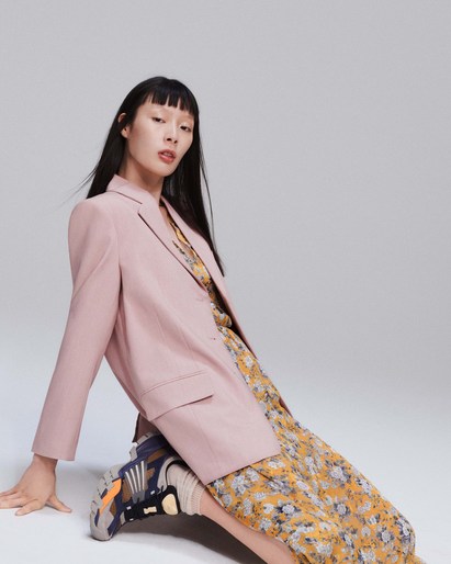 Asian woman in a pink blazer and yellow floral dress poses sitting on her sneakers against a neutral grey backdrop in a fashion studio.